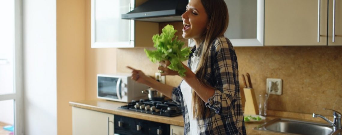 Cheerful young funny woman dancing and singing with lettuce microphone while cooking breakfast in the kitchen early morning at home